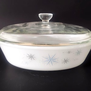 May include: White oval casserole dish with a clear glass lid. The dish features a starburst pattern in blue and black. The lid has a clear glass knob. The dish has a gold rim.