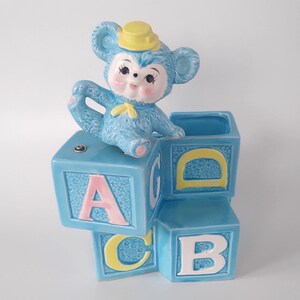 May include: A blue ceramic baby block planter with a blue monkey figurine wearing a yellow hat. The blocks are arranged in a stack and feature the letters A, B, C, and D in pink and yellow.