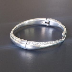 May include: A silver-toned hinged bangle bracelet featuring a smooth, polished finish on one side and a tapered section inlaid with a row of clear, sparkling crystals. The bracelet includes a secure metal hinge mechanism and a small decorative loop.