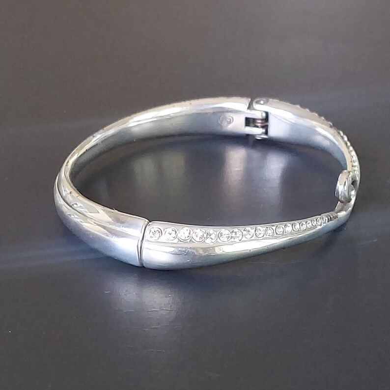 May include: A silver-toned hinged bangle bracelet featuring a smooth, polished finish on one side and a tapered section inlaid with a row of clear, sparkling crystals. The bracelet includes a secure metal hinge mechanism and a small decorative loop.
