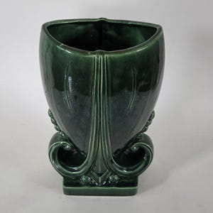 May include: A dark green ceramic vase with a glossy finish, featuring a wide, flared top opening and a structured base with decorative scrollwork and small spherical accents on the sides. The body has vertical molded lines and a square base.