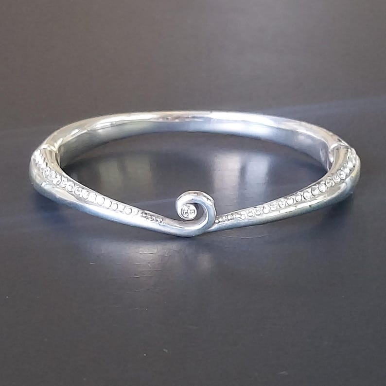 May include: A silver-toned hinged bangle bracelet featuring a central spiral design element. The surface of the bracelet is embellished with a row of small, clear, sparkling stones along the sides, leading toward the prominent center swirl.