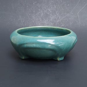 May include: A teal ceramic bowl with a smooth, glossy finish and a rounded, low-profile shape. The bowl features a wide, open top and is supported by three small, integrated feet at the base, creating a stable and balanced appearance.