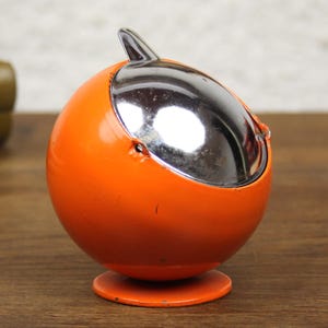 May include: A spherical orange metal ashtray featuring a polished chrome rotating lid with a small pointed handle. The round container rests on a circular orange base, showcasing a smooth, glossy finish and a compact, functional design.