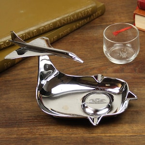 May include: A polished chrome metal tray featuring a three-dimensional model of a supersonic airplane attached to the side. The tray has a smooth, reflective surface with a circular indentation in the center containing a small embossed airplane logo.
