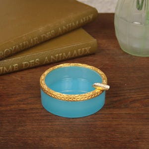 May include: A round, light blue glass ashtray featuring a textured, metallic gold-toned rim. The ashtray has a single small notch on the side for resting a cigarette, and the overall design is simple, circular, and compact in its construction.