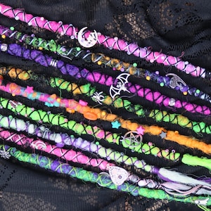 May include: A selection of vibrant dreadlock extensions in shades of pink, purple, green, and orange. Each dreadlock is decorated with charms, including moons, stars, and spiderwebs. The extensions are displayed on a black lace background.