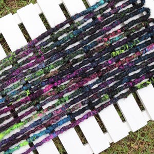May include: A selection of multi-coloured dreadlocks, predominantly black with streaks of purple, green, and blue, displayed on a white slatted surface. These handmade dreadlocks offer a vibrant and unique aesthetic for various hairstyles.