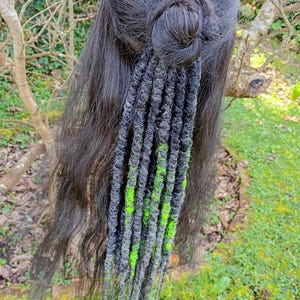 May include: Black hair styled with a bun and accented with gray and neon green dreadlocks. The dreadlocks are attached to the hair and hang down. The background is a blurred view of greenery.