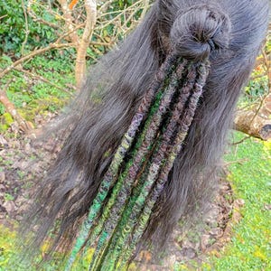 May include: Dark hair styled in a bun with several green, brown, and purple dreadlock extensions. The extensions are attached to the back of the head, cascading down. The image includes the text "Black Sunshine" in a banner.