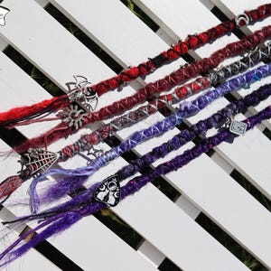 May include: A selection of vibrant dreadlocks in red, purple, and blue, embellished with metal charms. The dreadlocks are displayed on a white slatted surface, with the text "Black Sunshine" visible in the upper left corner.