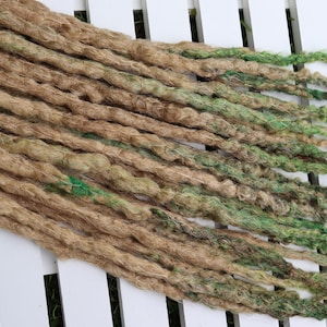 May include: A set of synthetic dreadlocks featuring a blend of natural tan and light green fibers. The textured, twisted strands vary in thickness and length, creating a layered appearance. A small white tag with the text Black Sunshine is visible.