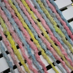 May include: A collection of pastel-coloured dreadlocks in pink, blue, and yellow, arranged diagonally on a white slatted surface. The dreadlocks appear thick and textured, with a handmade look. The Black Sunshine logo is visible.