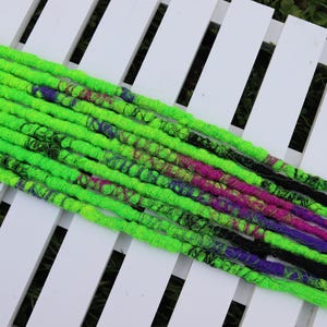 May include: Vibrant neon green dreadlocks with streaks of pink, purple, and black. The dreads are arranged on a white slatted surface. These dreadlocks are a striking accessory for alternative hairstyles.