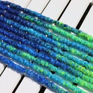 May include: Close-up of several blue, teal, and green dreadlocks. The dreadlocks are arranged diagonally across a white surface. The color transitions from dark blue to teal and then to green. The Black Sunshine logo is in the top right corner.