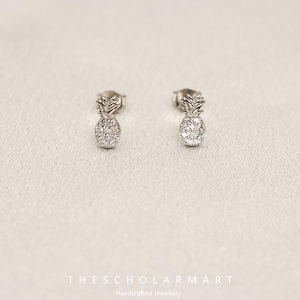 Puede incluir: Un par de pendientes de botón plateados con forma de piñas en miniatura. Los cuerpos de las piñas están incrustados con pequeñas piedras brillantes. Los pendientes se muestran sobre una superficie blanca texturizada.