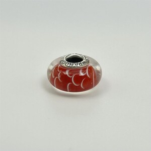 May include: A red and clear glass bead with white crescent-shaped patterns. The bead features a silver-coloured metal core with the word "PANDORA" engraved. A decorative charm.