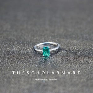 Peut inclure: Une bague en argent ornée d'une pierre verte rectangulaire taille émeraude. La bague est posée sur une surface texturée gris foncé. Le texte "THESCHOLARMART" et "Handcrafted Jeweller" sont visibles.
