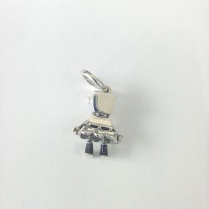 May include: A silver charm in the shape of a doll. The charm has a rectangular head, a scalloped skirt, and two black legs. The charm is suspended from a silver loop.