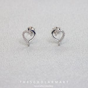 Puede incluir: Un par de pendientes de botón plateados con forma de corazón. Cada pendiente presenta un contorno de corazón con una fila de pequeñas piedras brillantes a lo largo de la curva superior. Los pendientes se muestran sobre un fondo gris claro. El texto "THESCHOLARMART Handcrafted Jewellery" está en la parte inferior.