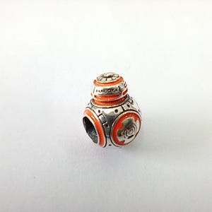 May include: A silver charm with orange and white enamel detailing. The charm is spherical with a domed top and features a design reminiscent of a droid from a popular science fiction franchise. The word "PANDORA" is visible on the top.