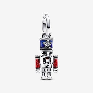 May include: A silver nutcracker charm featuring a blue enamel hat with a central star and a clear stone, black enamel eyes and mustache details, and red glitter enamel accents on the arms. The charm includes a circular bail for attachment.
