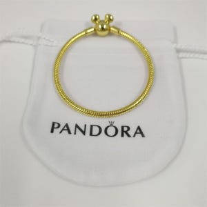 Pode incluir: Uma pulseira dourada com um fecho em forma de Mickey Mouse repousa sobre uma bolsa branca. A pulseira tem um design texturizado em forma de cobra. A bolsa tem a palavra "PANDORA" impressa em preto.