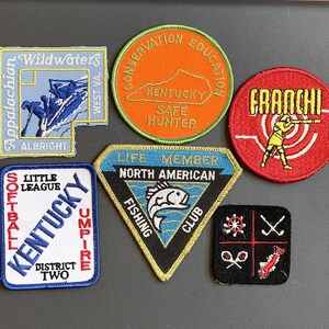 May include: A collection of six embroidered patches including a blue Appalachian Wildwaters patch, an orange Kentucky Conservation Education patch, a red Franchi target patch, a white Kentucky Little League Umpire patch, a triangular North American Fishing Club patch, and a black square patch with four white icons.