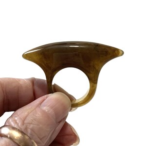 May include: A ring made of translucent brown resin with a marbled pattern. The ring features a wide, curved, horizontal top section that tapers to pointed ends, connected to a circular band for the finger, showcasing a smooth, polished finish.