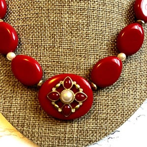 May include: A bold red necklace with large oval beads and a central pendant. The pendant features a red oval base with a pearl and gold-toned accents. The necklace is displayed on a textured, neutral-toned surface.