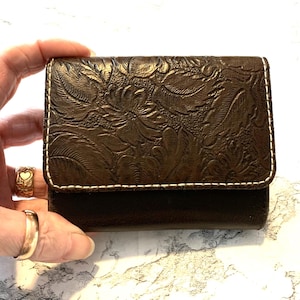May include: A brown leather wallet with a floral embossed design. The wallet has a rectangular shape with a white stitched border. The wallet is held in a hand.