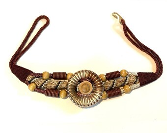 May include: A decorative belt with a central silver-toned, circular embellishment with a wooden centre. The belt has a burgundy braided cord and is accented with wooden beads and woven fabric. The belt is designed with a hook closure.