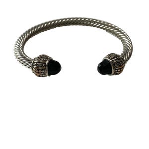 May include: A silver-toned cuff bracelet featuring a twisted cable design. The two ends of the bracelet are capped with textured metal ornaments, each set with a smooth, black, pointed gemstone that faces inward toward the center opening.