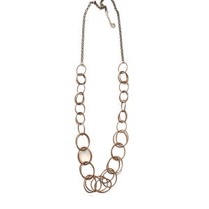May include: A long necklace featuring a series of interconnected, bronze-colored rings of varying sizes. The necklace has a silver-toned chain and clasp. The rings are arranged in a cascading pattern, creating a layered effect.
