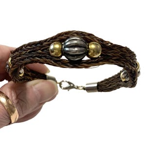 May include: A bracelet crafted from braided brown hair, featuring a central ribbed silver-toned bead flanked by two smooth gold-toned beads. The band includes additional alternating silver and gold-toned metal accents and a silver-toned lobster clasp.