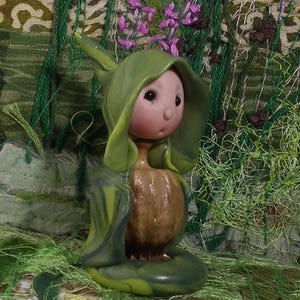 May include: A small figurine featuring a round, pale pink face with dark eyes and a small nose, wearing a hooded green cloak. The body is shaped like a textured, brown, bulbous root or gourd, standing on a smooth, rounded green base.