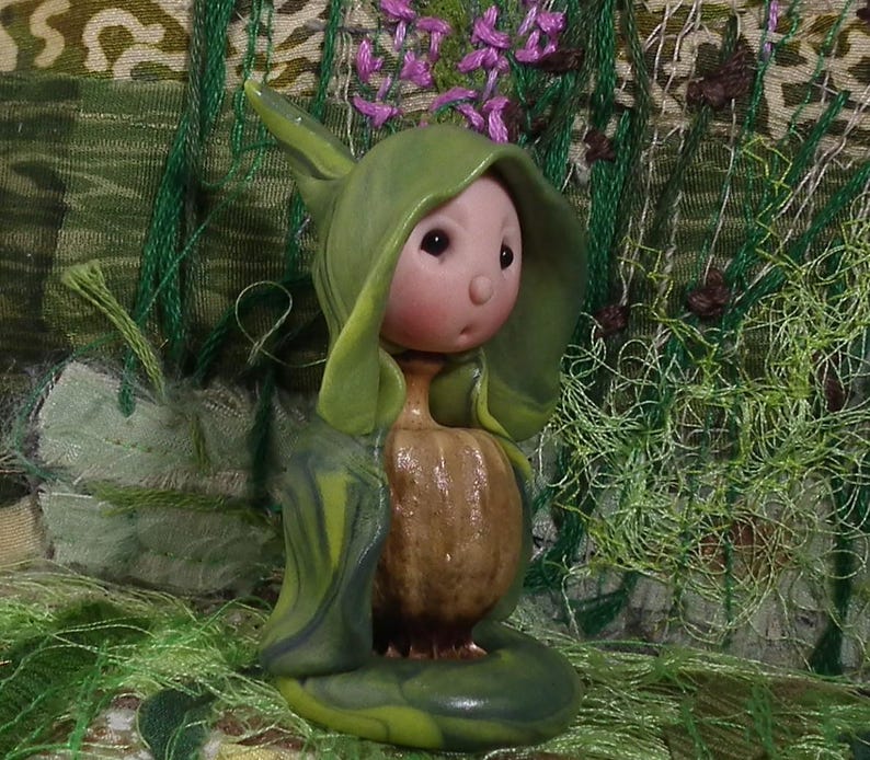 May include: A small figurine featuring a round, pale pink face with dark eyes and a small nose, wearing a hooded green cloak. The body is shaped like a textured, brown, bulbous root or gourd, standing on a smooth, rounded green base.