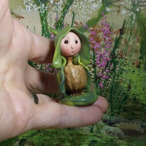 May include: A small figurine featuring a pale, round-faced character with rosy cheeks, wearing a green hooded cloak that drapes over a textured, brown, bulbous body. The character has a small nose and a tiny, open mouth, with a smooth finish.