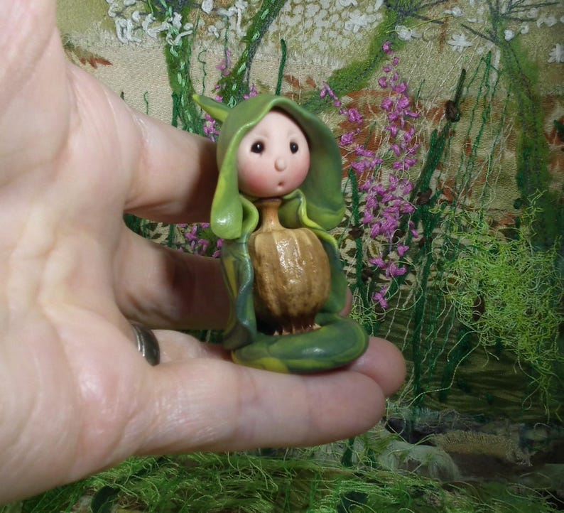 May include: A small figurine featuring a pale, round-faced character with rosy cheeks, wearing a green hooded cloak that drapes over a textured, brown, bulbous body. The character has a small nose and a tiny, open mouth, with a smooth finish.