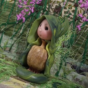 May include: A small figurine featuring a pale, round-faced character with a tiny nose and dark eyes, wearing a hooded green cloak with swirling patterns. The character holds a textured, tan, pumpkin-shaped object in front of its body.