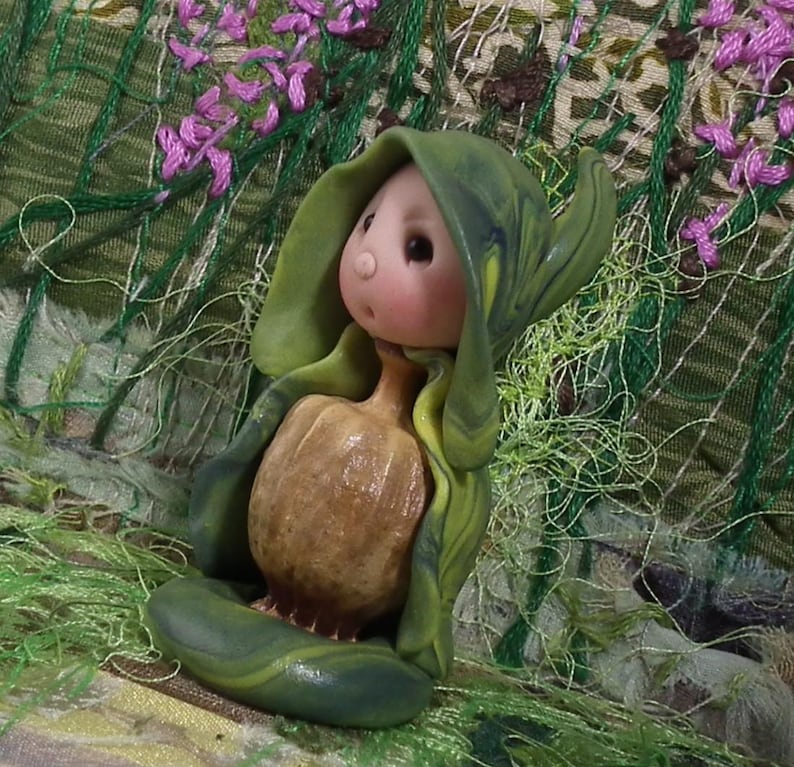 May include: A small figurine featuring a pale, round-faced character with a tiny nose and dark eyes, wearing a hooded green cloak with swirling patterns. The character holds a textured, tan, pumpkin-shaped object in front of its body.