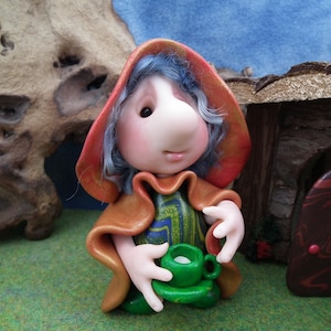 Op de afbeelding: Een grillig beeldje van een zittend personage met een grote neus, blauw haar en een rode capuchon. De figuur houdt een groene kop en schotel vast. Het personage draagt een groen en blauw patroon tuniek. De achtergrond omvat een houten structuur en groen gras.