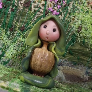 Könnte beinhalten: Eine kleine Figur mit einem runden, blassrosa Gesicht, dunklen Augen und einem kleinen Mund, die einen grünen Kapuzenumhang über einem strukturierten, beigefarbenen, knolligen Körper trägt. Die Figur sitzt auf einem glatten, dunkelgrünen Sockel.