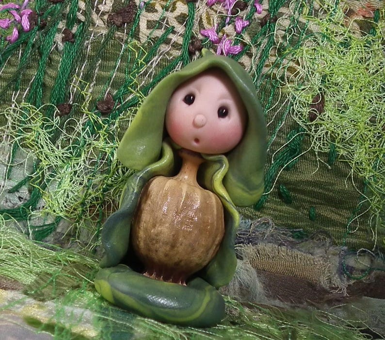 May include: A small figurine featuring a round, pale pink face with dark eyes and a small mouth, wearing a green hooded cloak that drapes over a textured, tan, bulbous body. The figure sits on a smooth, dark green base with a rounded shape.