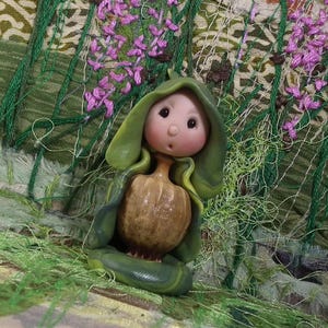 May include: A small figurine featuring a round, pale-skinned face with dark eyes and a small mouth, emerging from a brown, textured, pumpkin-shaped body. The figure is draped in a smooth, layered green hood that wraps around the torso.