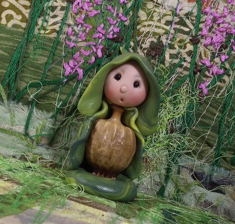 May include: A small figurine featuring a round, pale-skinned face with dark eyes and a small mouth, emerging from a brown, textured, pumpkin-shaped body. The figure is draped in a smooth, layered green hood that wraps around the torso.