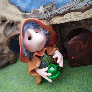 May include: A whimsical figurine of a gnome-like character with blue hair, wearing a red hood and brown cloak, holding a green teacup and saucer. The miniature scene includes a wooden structure and a grassy area.