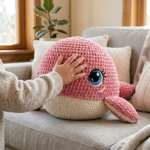 May include: A plush crochet whale toy featuring a textured pink upper body and a cream-colored underbelly. The whale has large, embroidered blue eyes with black pupils and long eyelashes, along with a small stitched white mouth and side fins.
