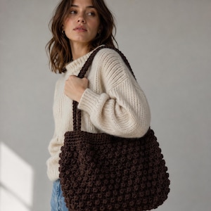 May include: A dark brown crochet tote bag featuring a textured bobble stitch pattern throughout the body and two thick, matching crocheted shoulder straps. The bag is constructed from yarn and has a rectangular shape with a wide, open top opening.