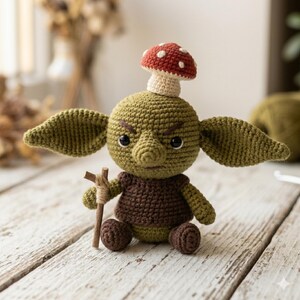 May include: A crocheted green goblin doll with large pointed ears, black eyes, and a stern expression. The doll wears a brown tunic, has a red and white mushroom cap on its head, and holds a small wooden staff in its right hand.